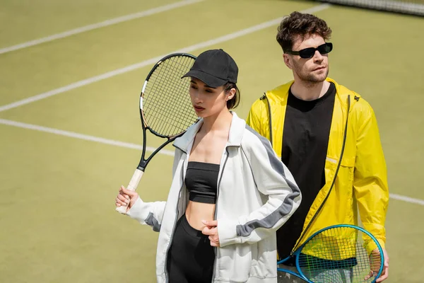Модная спортивная пара, мужчина в солнцезащитных очках и женщина в кепке, держащая ракетки на теннисном корте — стоковое фото