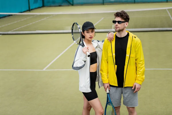 Estilo e esporte, casal atlético, homem de óculos de sol e mulher de boné segurando raquetes no campo de ténis — Fotografia de Stock