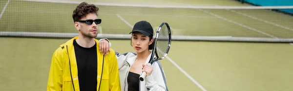 Pancarta, estilo y deporte, hombre atlético en gafas de sol y mujer en gorra sosteniendo raqueta en pista de tenis - foto de stock