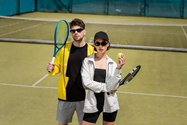 Moda y deporte, hombre en gafas de sol y mujer en gorra sosteniendo raquetas y pelota en pista de tenis - foto de stock