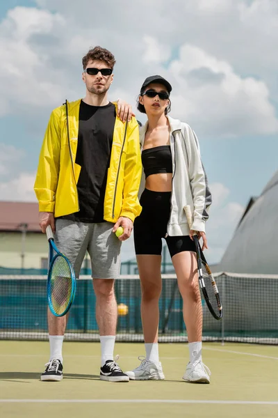 Hobby e sport, uomo e donna alla moda in occhiali da sole con racchette e palla sul campo da tennis — Foto stock