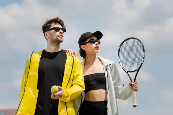 Tenis deporte y moda, hombre y mujer en gafas de sol con raqueta y pelota en la cancha de tenis, hobby - foto de stock