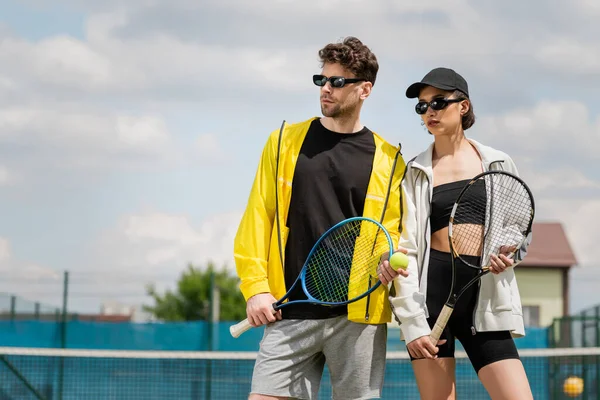 Deporte de verano, hombre y mujer en gafas de sol de pie en la cancha de tenis con raquetas, los atletas de moda - foto de stock