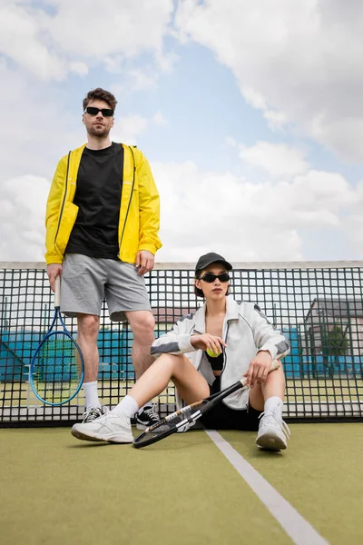 Pareja deportiva, hombre y mujer en gafas de sol posando en pista de tenis con raquetas, moda de deportistas - foto de stock