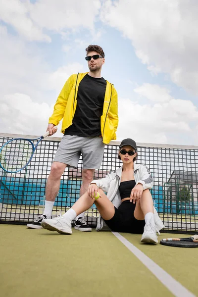 Pareja deportiva, hombre y mujer en gafas de sol posando cerca de la red de tenis con raquetas, moda deportiva - foto de stock