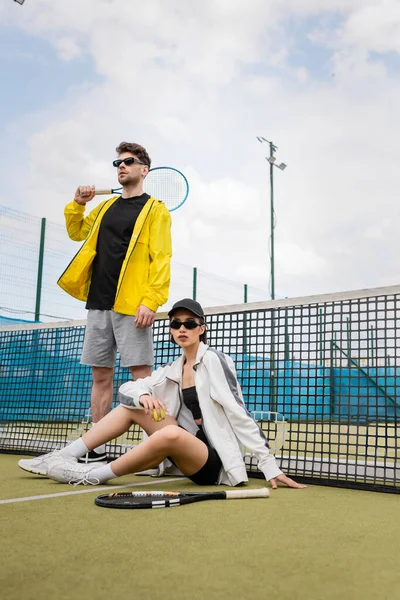 Хобби и спорт, мужчина и женщина в солнцезащитных очках позируют возле теннисной сетки с ракетками, спортивная мода — стоковое фото