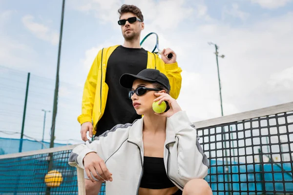Donna in occhiali da sole e cappello in posa vicino all'uomo atletico con racchetta da tennis, moda abbigliamento attivo — Foto stock