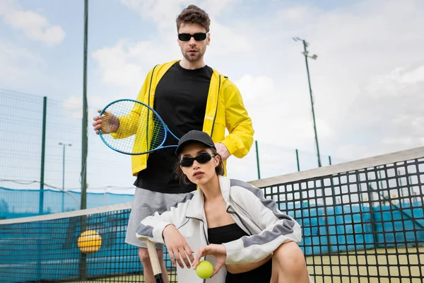 Mujer en gafas de sol y gorra posando cerca de hombre, sosteniendo pelota y raqueta de tenis, moda de desgaste activo - foto de stock