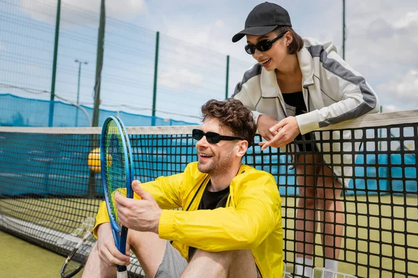 Hombre y mujer felices en ropa activa descansando cerca de la red de tenis en la cancha, ropa deportiva de moda, deporte - foto de stock