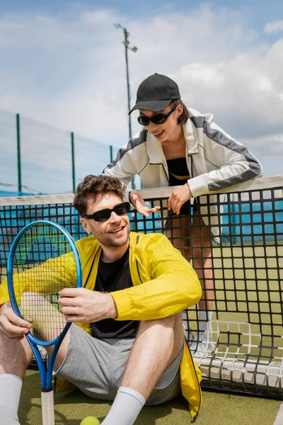 Mujer feliz en gafas de sol y ropa activa hablando con el hombre con raqueta de tenis, red de tenis, deporte - foto de stock