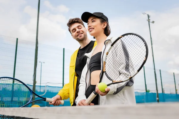 Cheerful couple in stylish active wear holding tennis rackets and ball on court, lifestyle and sport — Stock Photo