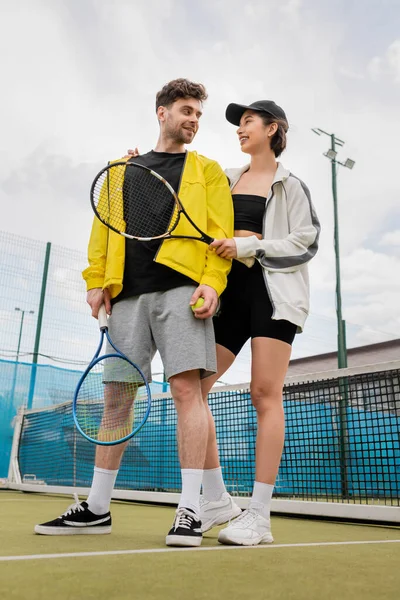 Donna felice in berretto e usura attiva tenendo racchetta e palla mentre abbraccia il fidanzato in campo, sport — Foto stock