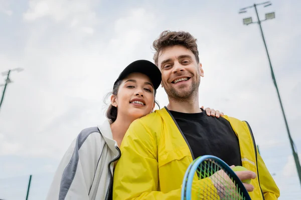 Mujer feliz en gorra y desgaste activo abrazo novio sosteniendo raqueta en la cancha, deporte, vista de bajo ángulo - foto de stock