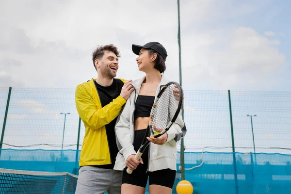 Позитивний чоловік в активному одязі обіймає жінку в шапці з тенісною ракеткою на корті, стилі життя і спорту — стокове фото
