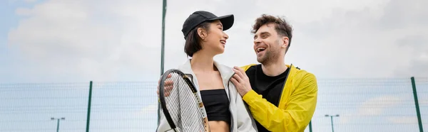 Банер, позитивний чоловік в активному одязі обіймає жінку в шапці з тенісною ракеткою на корті, стиль життя — стокове фото