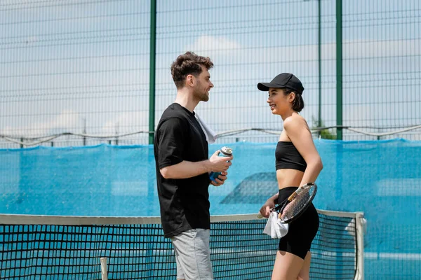 Homem feliz segurando garrafa de esportes com água perto de mulher com raquete de tênis na quadra, estilo de vida saudável — Fotografia de Stock