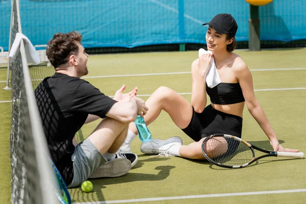 Feliz pareja charlando y sentado cerca de la red de tenis en la cancha, limpiando con toalla, raqueta y pelota - foto de stock