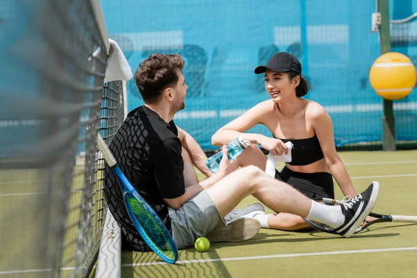 Feliz pareja en desgaste activo sentado con botella deportiva cerca de la red de tenis, raqueta de tenis y pelota, - foto de stock