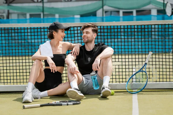 Щаслива пара відпочиває на тенісному корті, сидячи разом біля тенісної мережі, спортивної пляшки, ракетки, м'яча — стокове фото