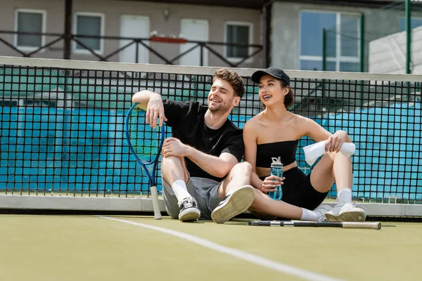Hombre y mujer felices en ropa deportiva descansando cerca de la red de tenis con raquetas en la cancha, estilo de vida saludable - foto de stock