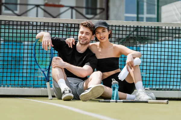 Hombre y mujer felices en ropa deportiva descansando cerca de la red de tenis en la cancha, pelota, raquetas, mirando a la cámara - foto de stock