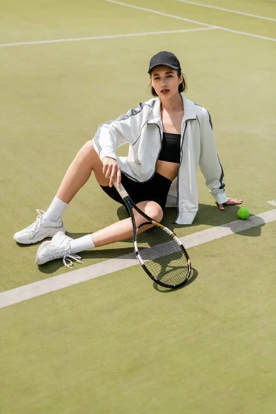 Mujer en desgaste activo y gorra sosteniendo raqueta de tenis y sentada en la cancha, jugadora de tenis - foto de stock
