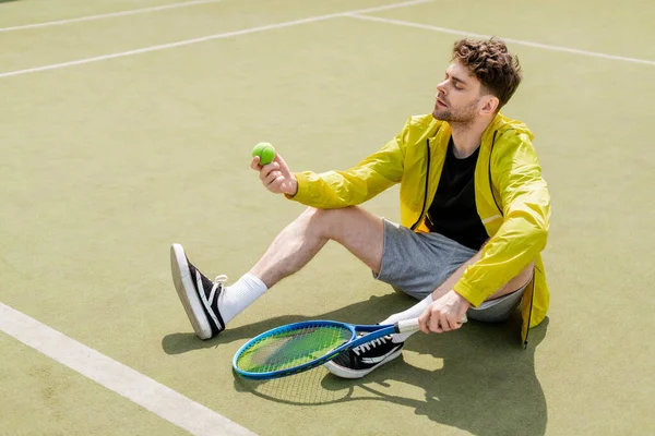 Joueur de tennis masculin assis sur le terrain et tenant la raquette, regardant la balle de tennis, homme en tenue active — Photo de stock