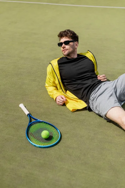 Homem bonito em óculos de sol descansando perto de bola de tênis e raquete, jogador de tênis masculino, esporte e estilo — Fotografia de Stock
