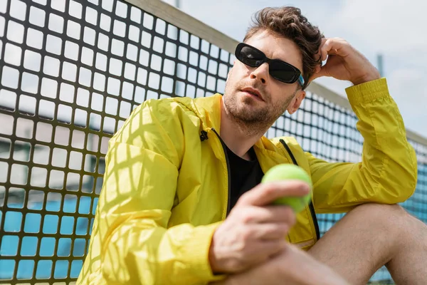 Athletic tennis player in sunglasses sitting near tennis net, holding ball, sport and style — Stock Photo