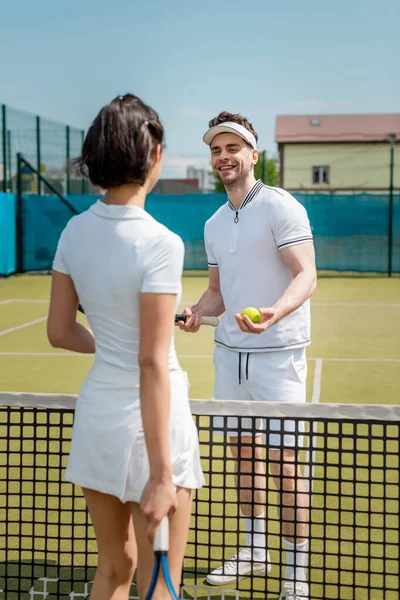 Hombre feliz mirando a la mujer cerca de la red de tenis, pareja de pie en la cancha de tenis, desgaste activo, pasatiempo - foto de stock