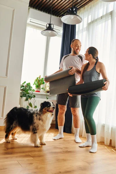 Positive couple in sportswear with fitness mats looking at each other near border collie at home — Stock Photo