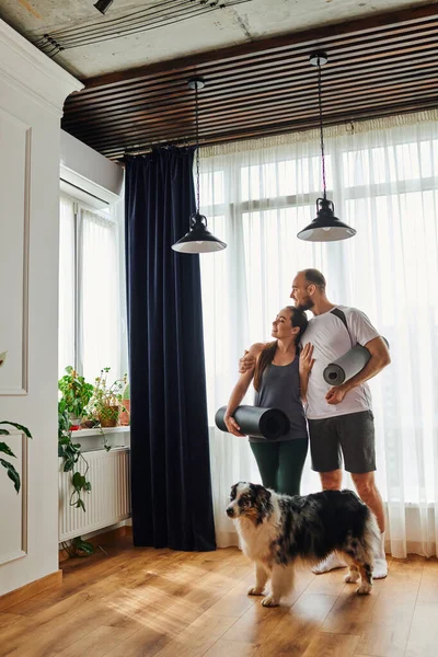 Smiling man in sportswear hugging girlfriend and holding fitness mat near border collie at home — Stock Photo