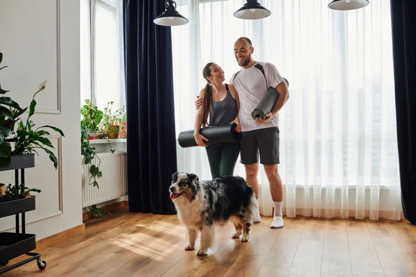 Un homme souriant en vêtements de sport étreignant sa petite amie avec un tapis de fitness et regardant border collie à la maison — Photo de stock
