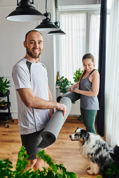 Homem feliz em sportswear olhando para a câmera perto da namorada com tapete de fitness e border collie em casa — Fotografia de Stock