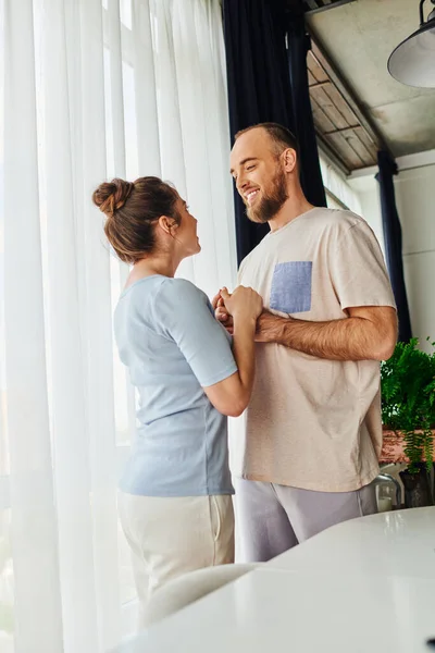 Pareja sonriente en ropa de casa tomados de la mano y mirándose mientras están de pie en casa - foto de stock