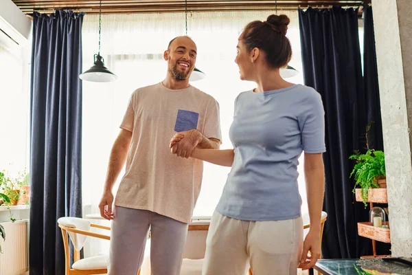 Smiling bearded man in homewear holding hand of girlfriend while spenging time together at home — Stock Photo