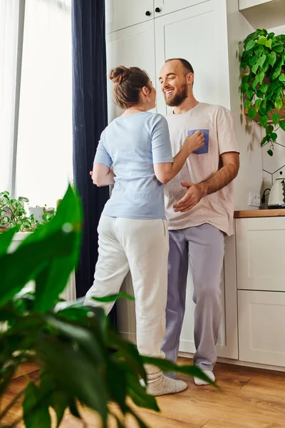 Joyful bearded man in loungewear talking to brunette girlfriend and standing in kitchen at home — Stock Photo