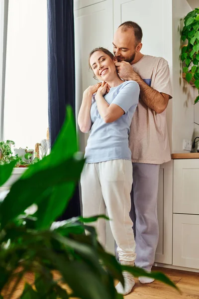 Bärtiger Mann in Homewear umarmt fröhliche brünette Freundin und steht zu Hause in der Küche — Stockfoto