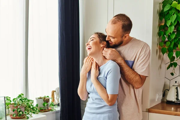 Positivo uomo barbuto in biancheria da casa abbracciando allegra fidanzata in cucina a casa — Foto stock