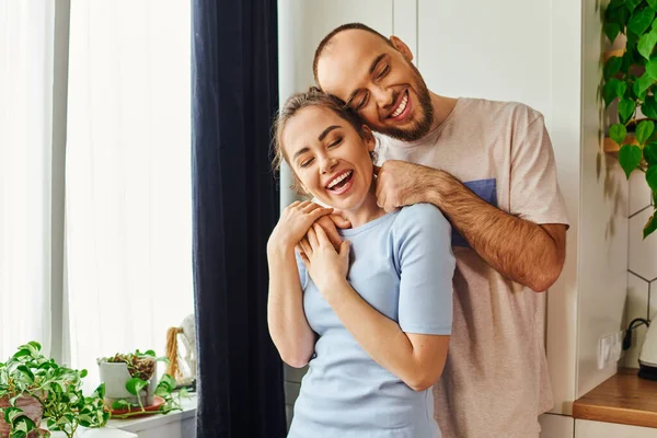 Joyful bearded man in homewear embracing girlfriend with closed eyes while standing at home — Stock Photo