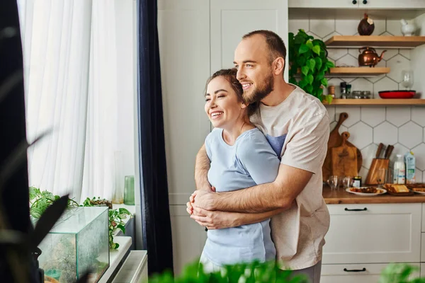 Positive man in homewear hugging brunette girlfriend while standing near plants and window at home — Stock Photo