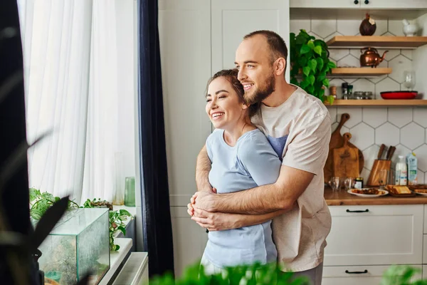 Hombre positivo en ropa de casa abrazando novia morena mientras está de pie cerca de las plantas y la ventana en casa - foto de stock