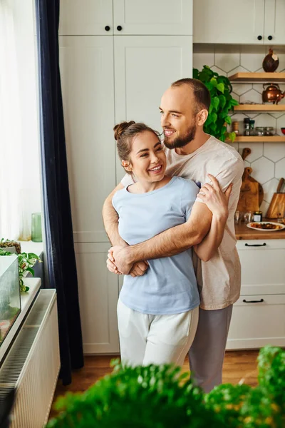 Sonriente hombre en ropa de casa abrazando novia morena mientras está de pie cerca de la ventana en la cocina en casa - foto de stock
