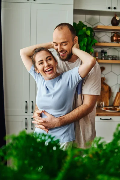Mujer morena sonriente en ropa de casa abrazando novio y de pie juntos en la cocina en casa - foto de stock