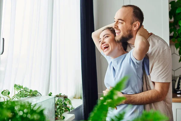 Joyful and relaxed couple in homewear hugging while standing near window at home — Stock Photo