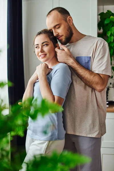 Tierno barbudo tocando a su novia sonriente en ropa de casa mientras pasan tiempo juntos en casa - foto de stock