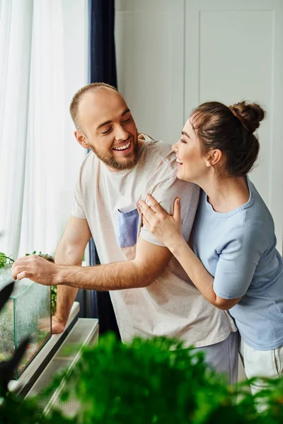 Smiling couple in homewear looking at each other while standing near terrarium at home — Stock Photo