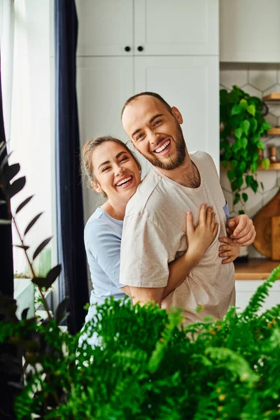 Smiling brunette woman in homewear hugging boyfriend and looking at camera near plants at home — Stock Photo