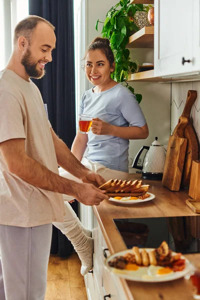 Улыбающаяся женщина, держащая чай, в то время как парень в домашней одежде берет тарелки с завтраком на кухне — стоковое фото