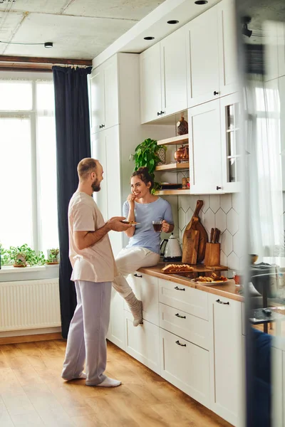 Femme positive en tenue de maison tenant le thé et regardant petit ami avec petit déjeuner dans la cuisine à la maison — Photo de stock